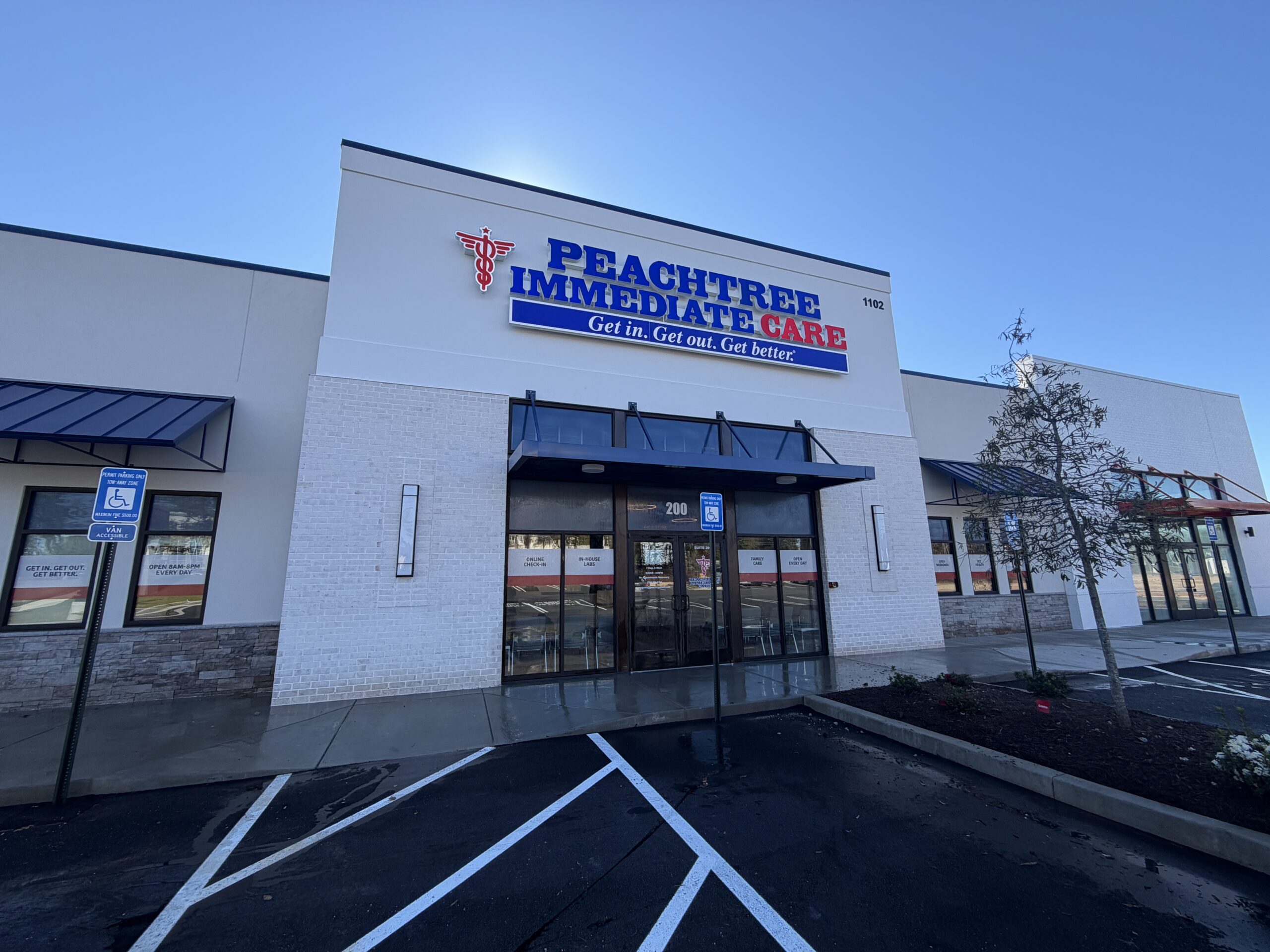 The entrance of Peachtree Immediate Care in Augusta, Georgia, on a bright, sun-filled day. A white building with a red medical caduceus, Peachtree Immediate in blue lettering and Care in red letting. With a smaller sign that reads "Get In. Get Out. Get Better." on a blue sign with white text underneath.
